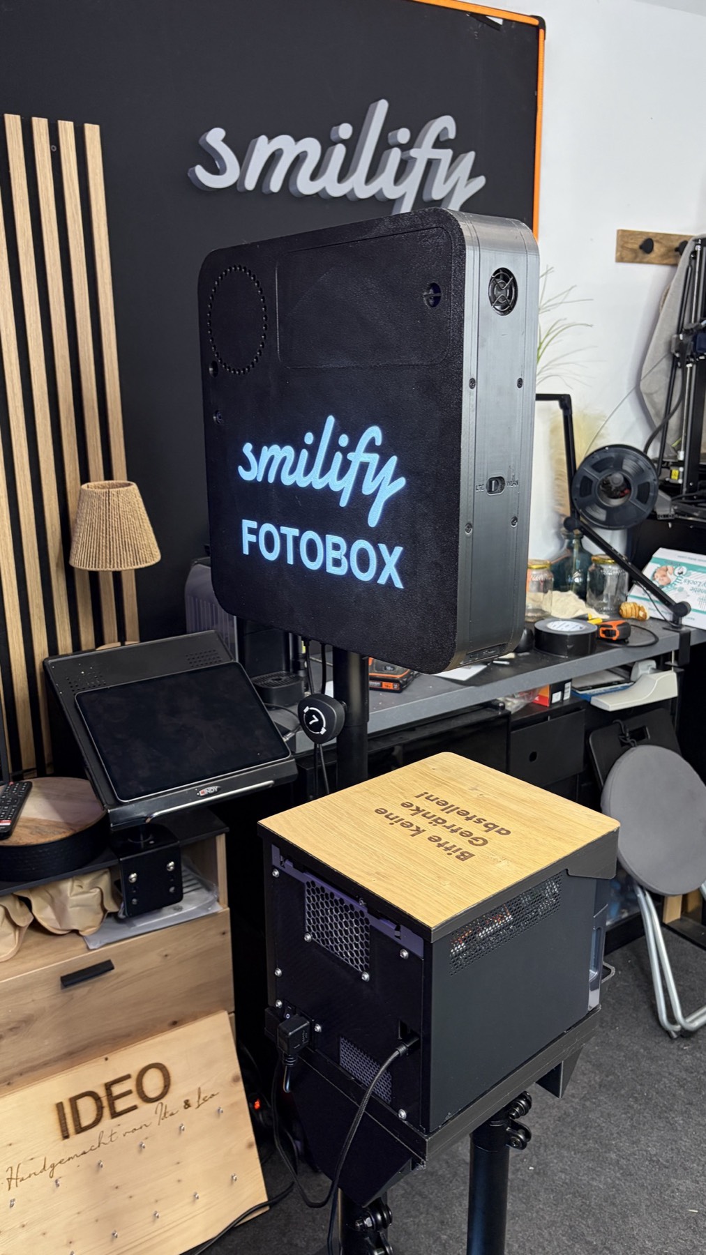 Die Smilify Fotobox von hinten mit leuchtendem Schriftzug smilify FOTOBOX, darunter auf dem Stativ das schwarze Druckergehäuse mit Holzplatte und der Aufschrift Bitte keine Getränke abstellen