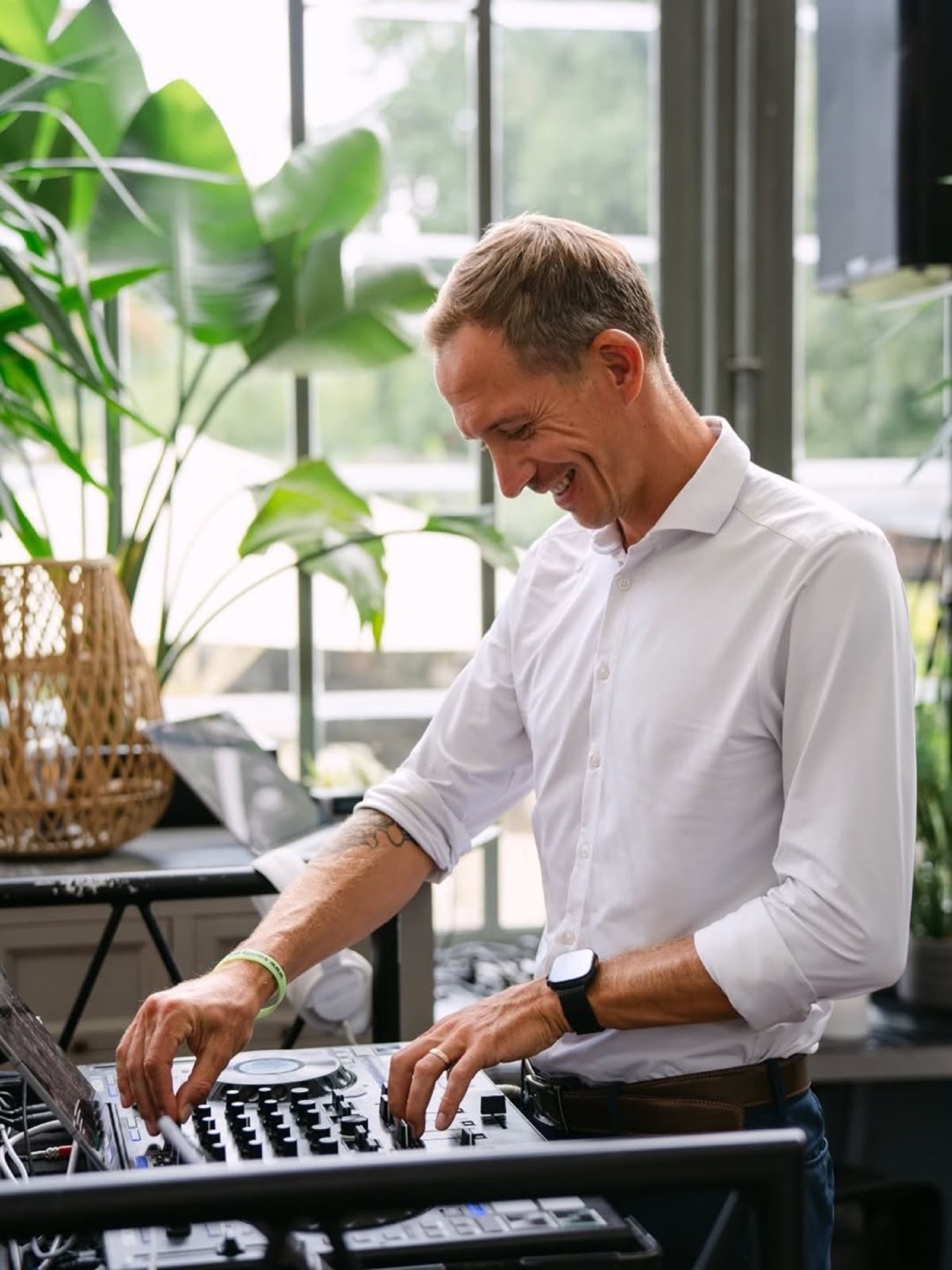 Chris lachend am DJ-Controller im hellen Schlosscafe im Palmenhaus, Pflanzen und Fenster im Hintergrund