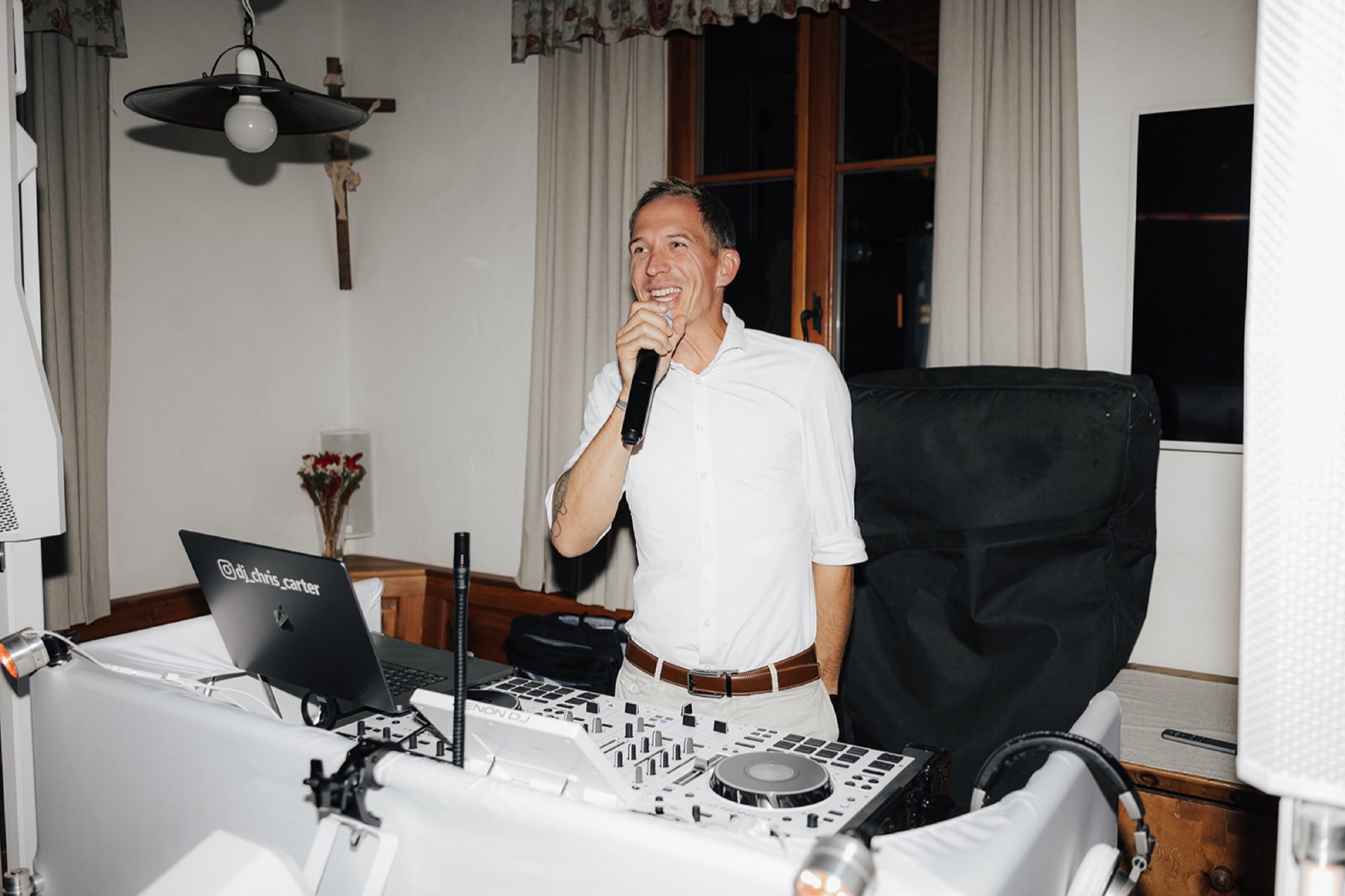 Chris moderiert lachend mit Mikro am weißen Pult, Laptop mit @dj_chris_carter, Stube mit Herrgottswinkel