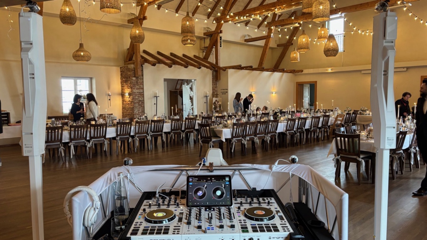 Blick über das weiße DJ-Pult in einen großen Saal mit Holzdecke und gedeckten Hochzeitstafeln