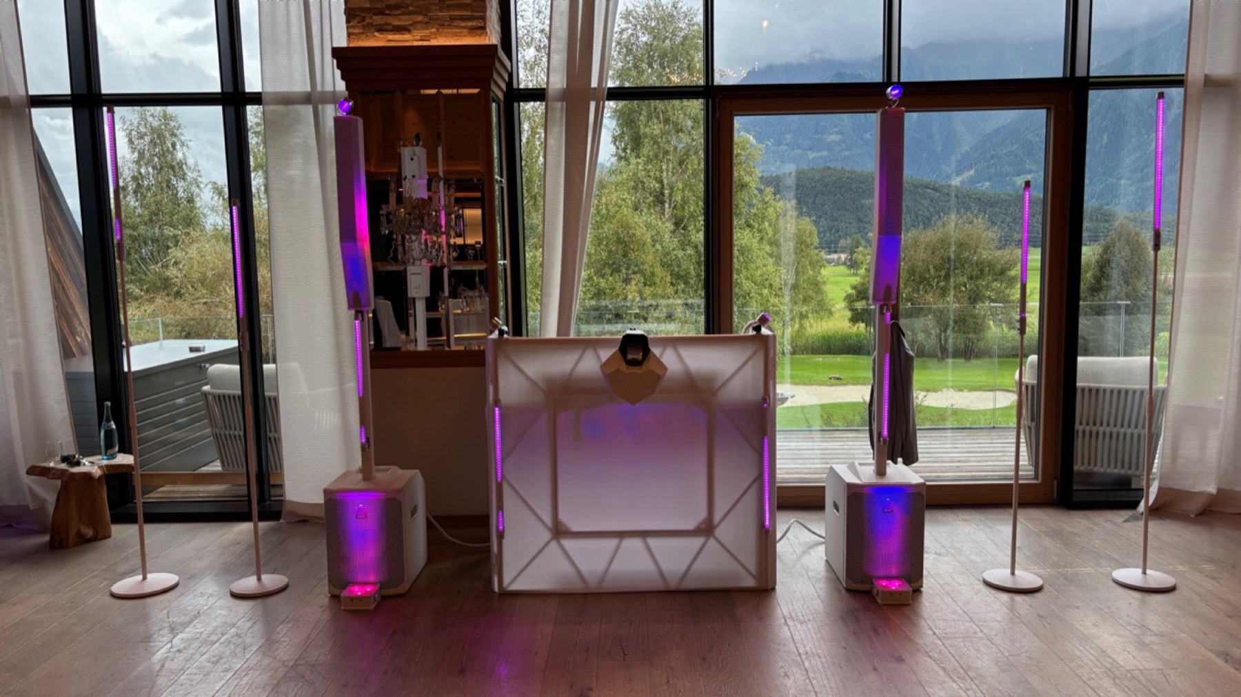 DJ-Pult mit Lautsprechern und violetten Lichtstangen vor einer Fensterfront mit Bergblick