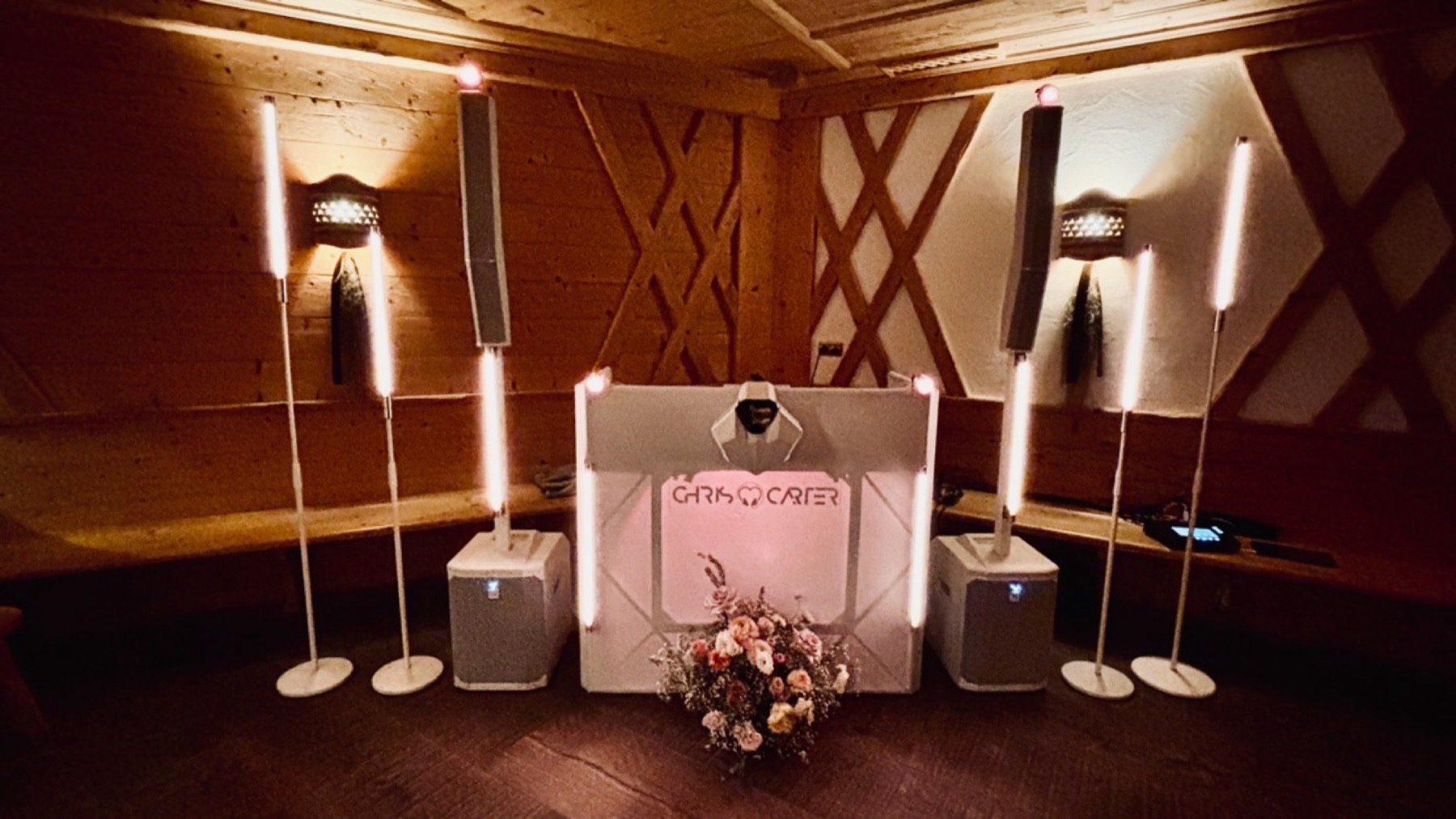 DJ-Pult mit Lautsprechern und Lichtstangen vor einer Holzwand mit Rautenmuster, warm beleuchtet