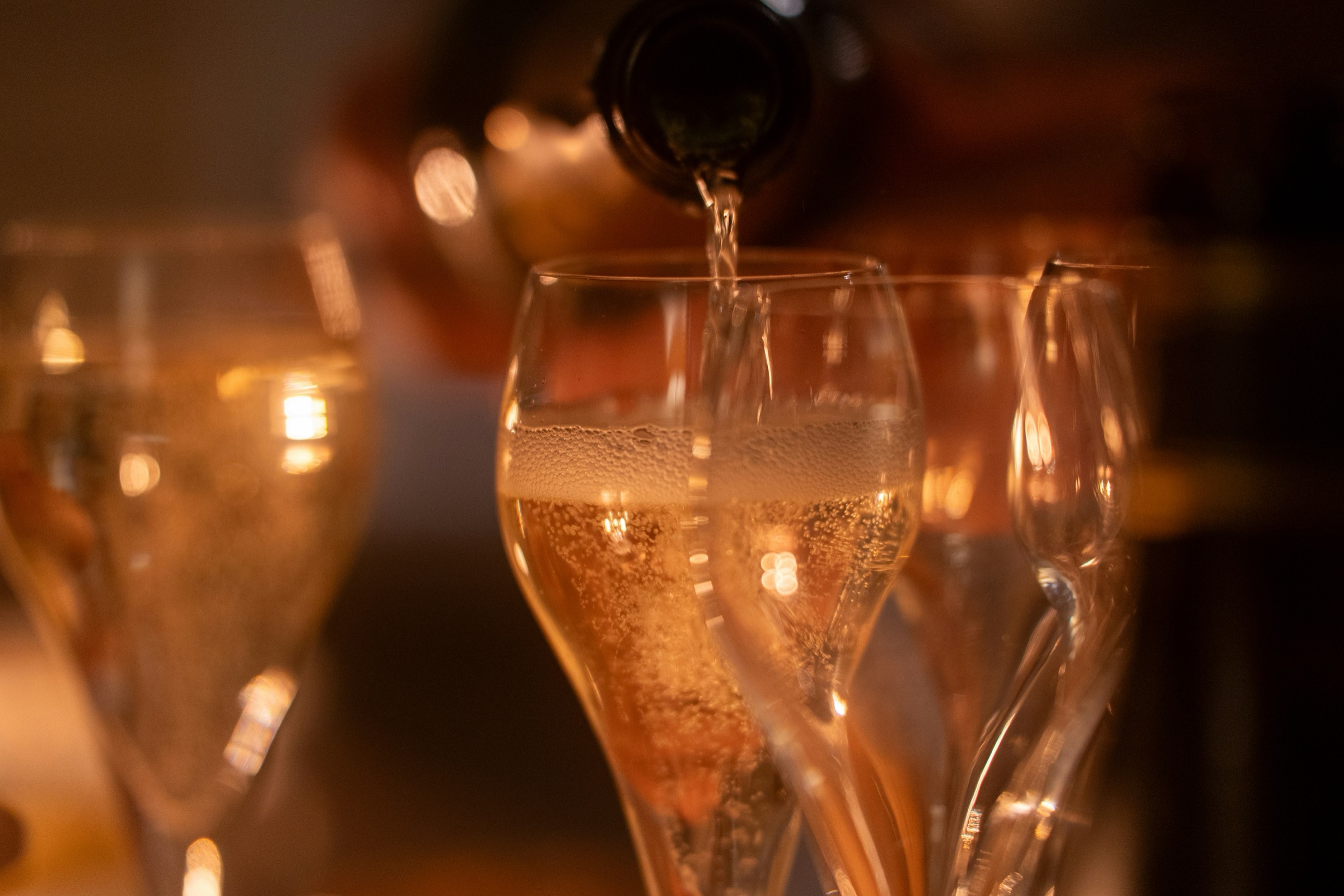 Sekt wird in schlanke Gläser eingeschenkt, warmes Licht im Hintergrund